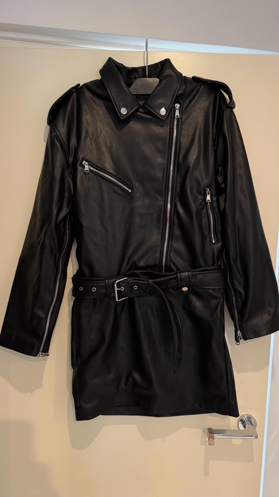 Black biker-style dress hanging on a door. It features a lapel collar, silver zips and a buckled belt.
