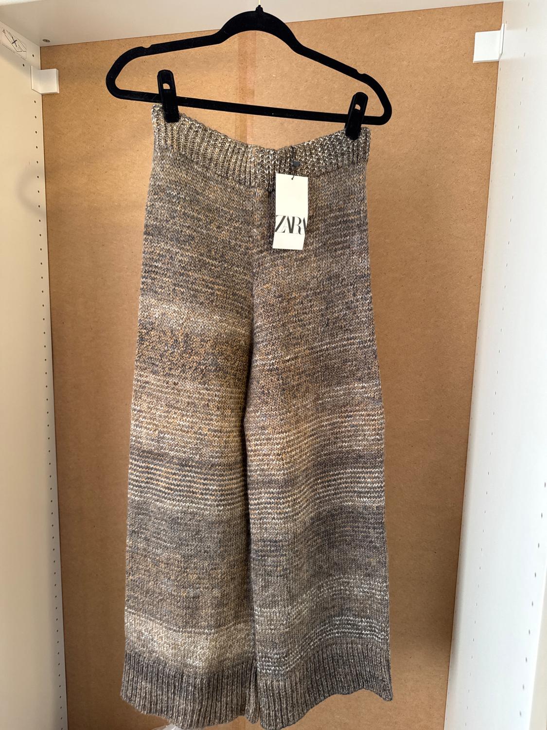 Grey and beige knitted trousers hanging on a black hanger with a visible "ZARA" label, light wood background.