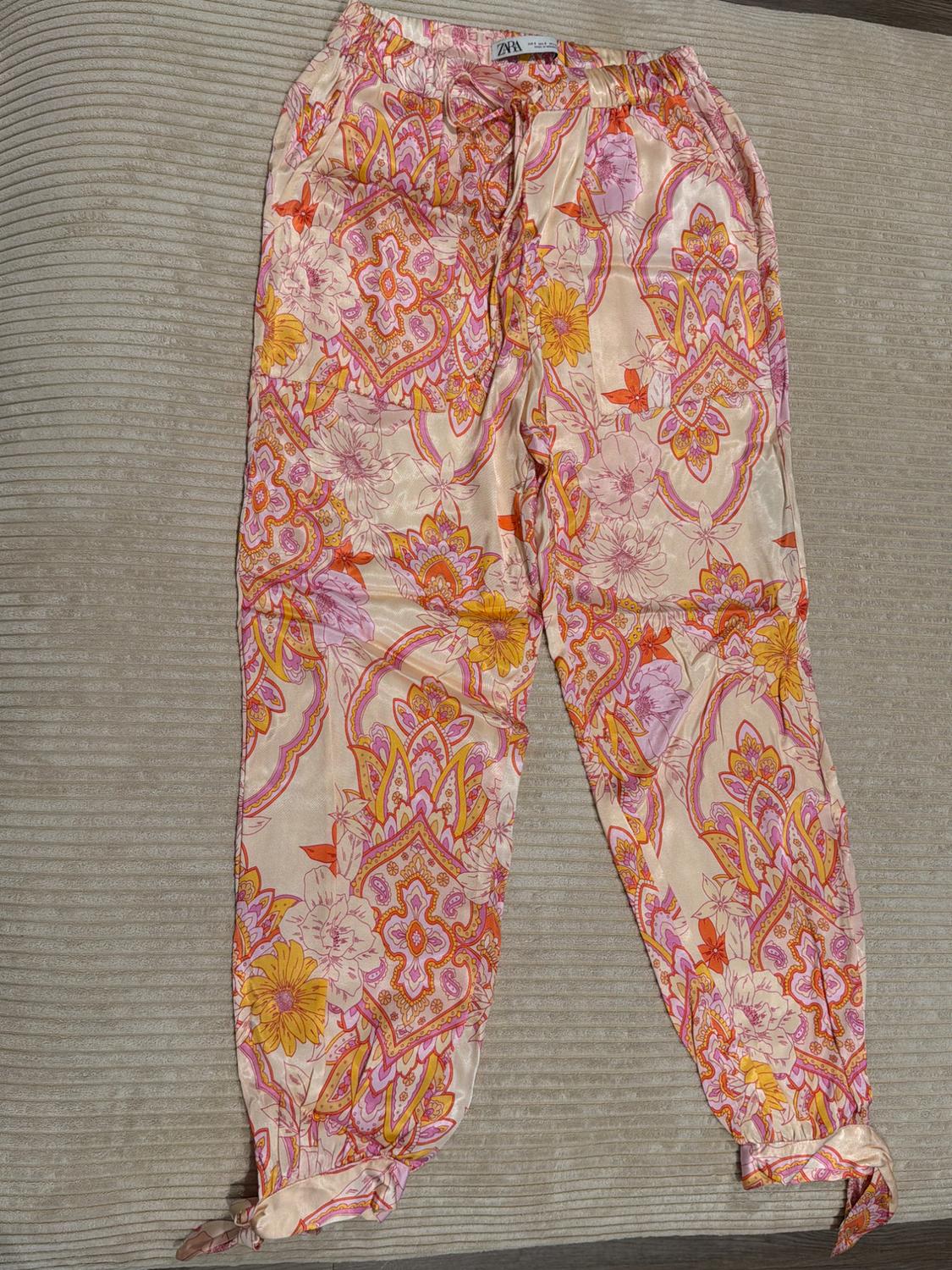 Pants with a pink, orange, and yellow floral print on a textured background. Elastic waistband with drawstring, knotted hems, and ZARA text.