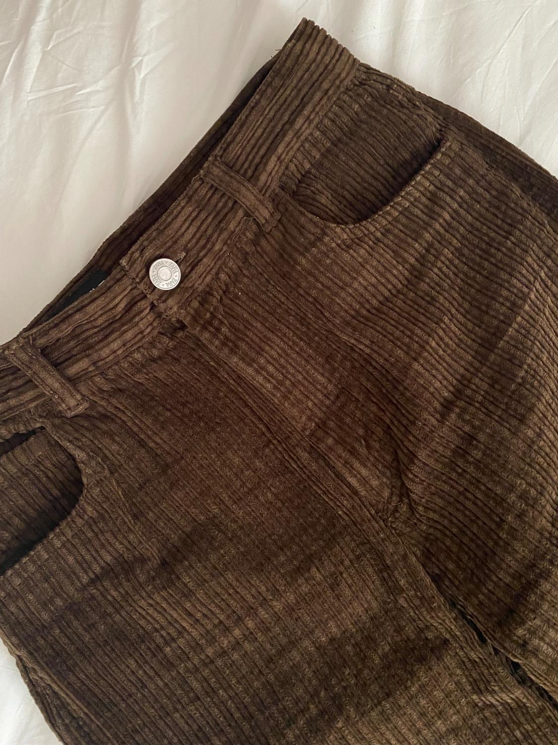 Close-up of a pair of brown corduroy trousers with pockets and a metal button that says ZARA, resting on a wrinkled white sheet.