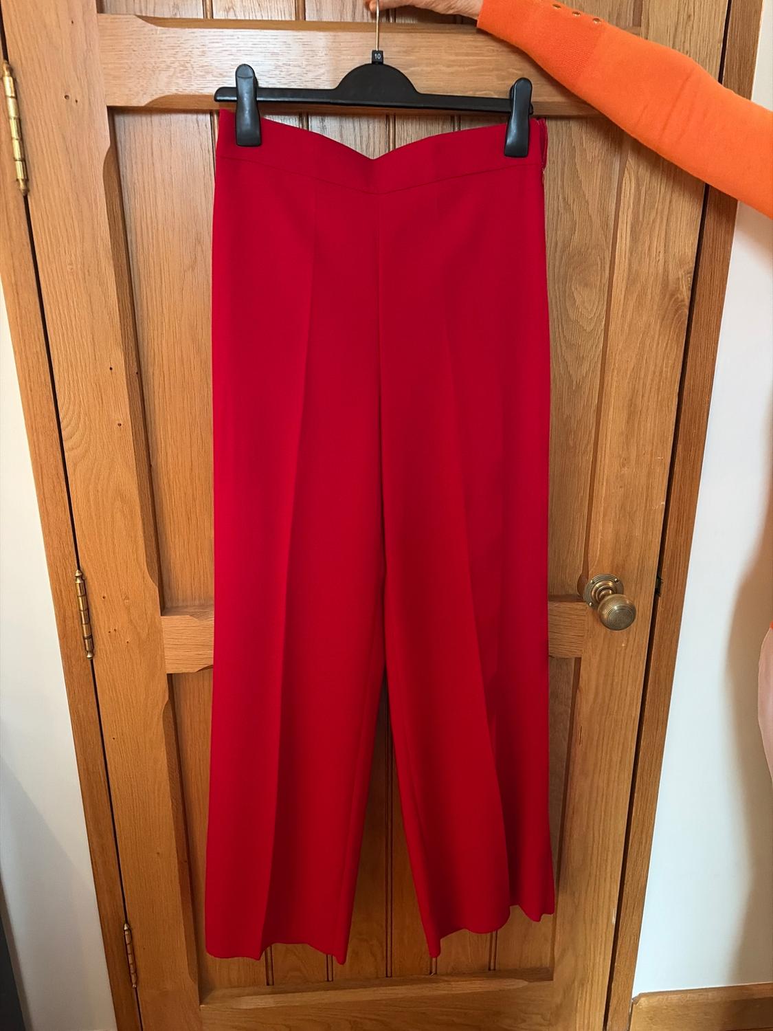 Red trousers are hanging on a black hanger over a wooden door. A hand wearing an orange jumper is holding the hanger.