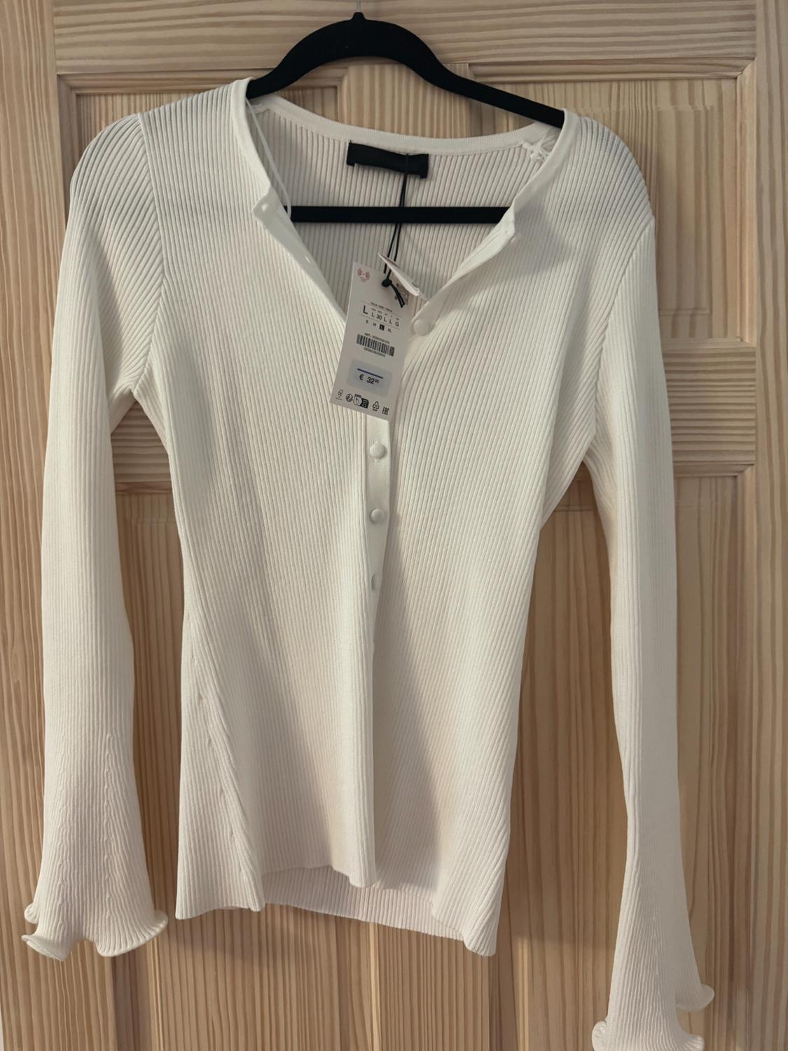 Ribbed white top with buttons and flared sleeves on a hanger over a wooden door. Text on tag: L €32.95.