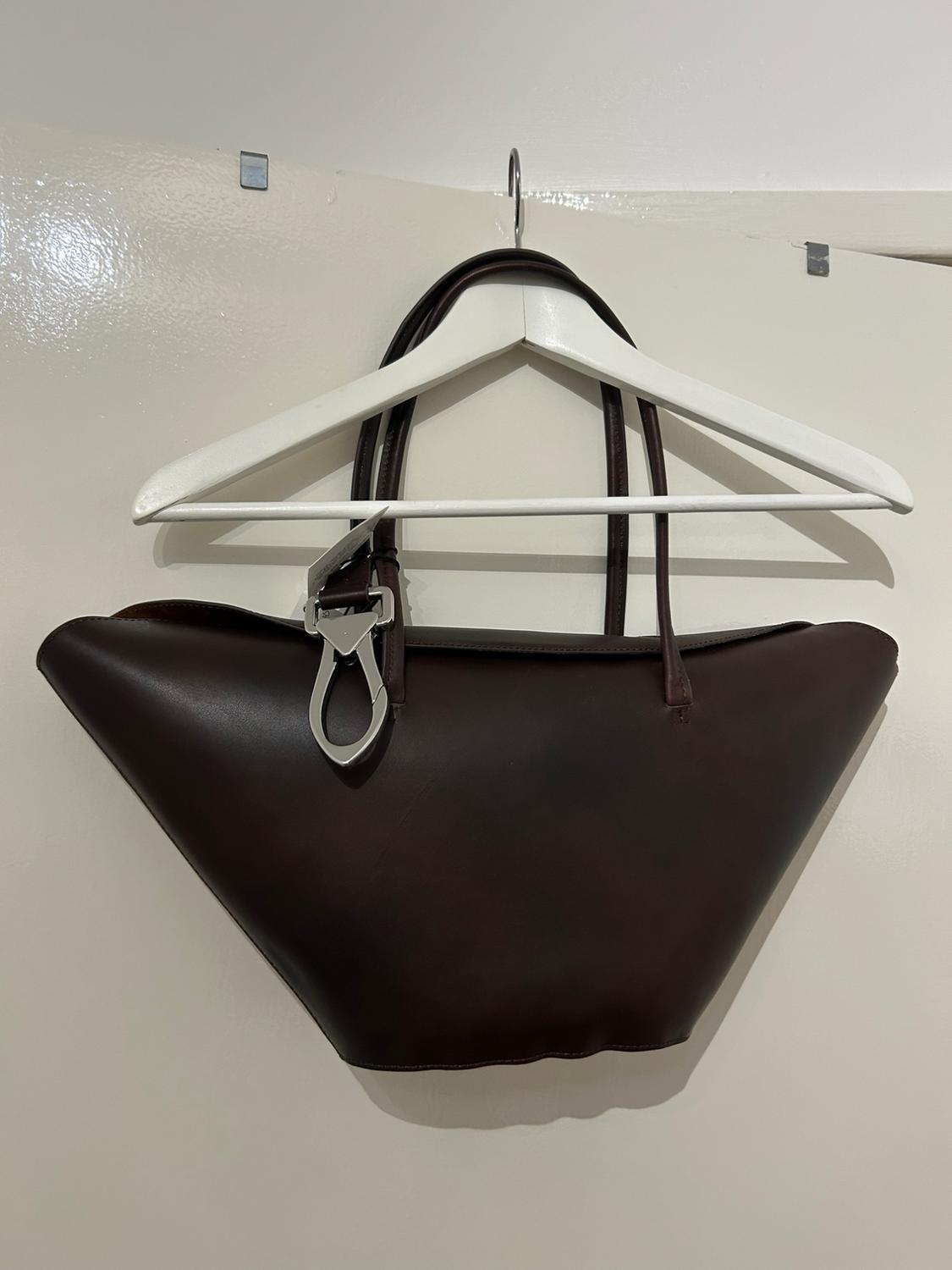 Dark brown handbag hanging on a white hanger against a light-coloured wall. The handbag has a decorative metal clasp.