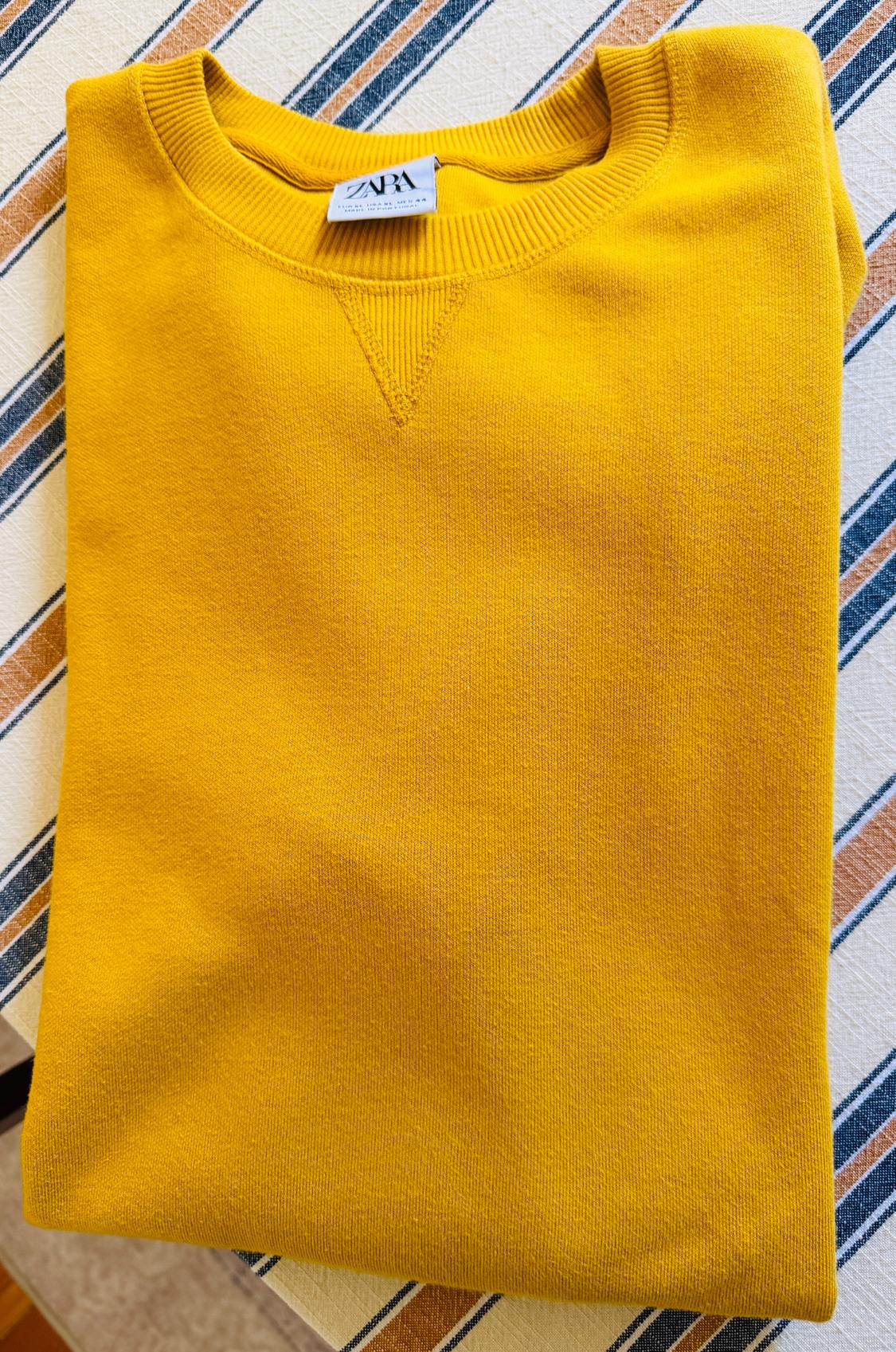 Yellow sweatshirt folded over a striped fabric. The round neck and the ZARA tag are prominent.