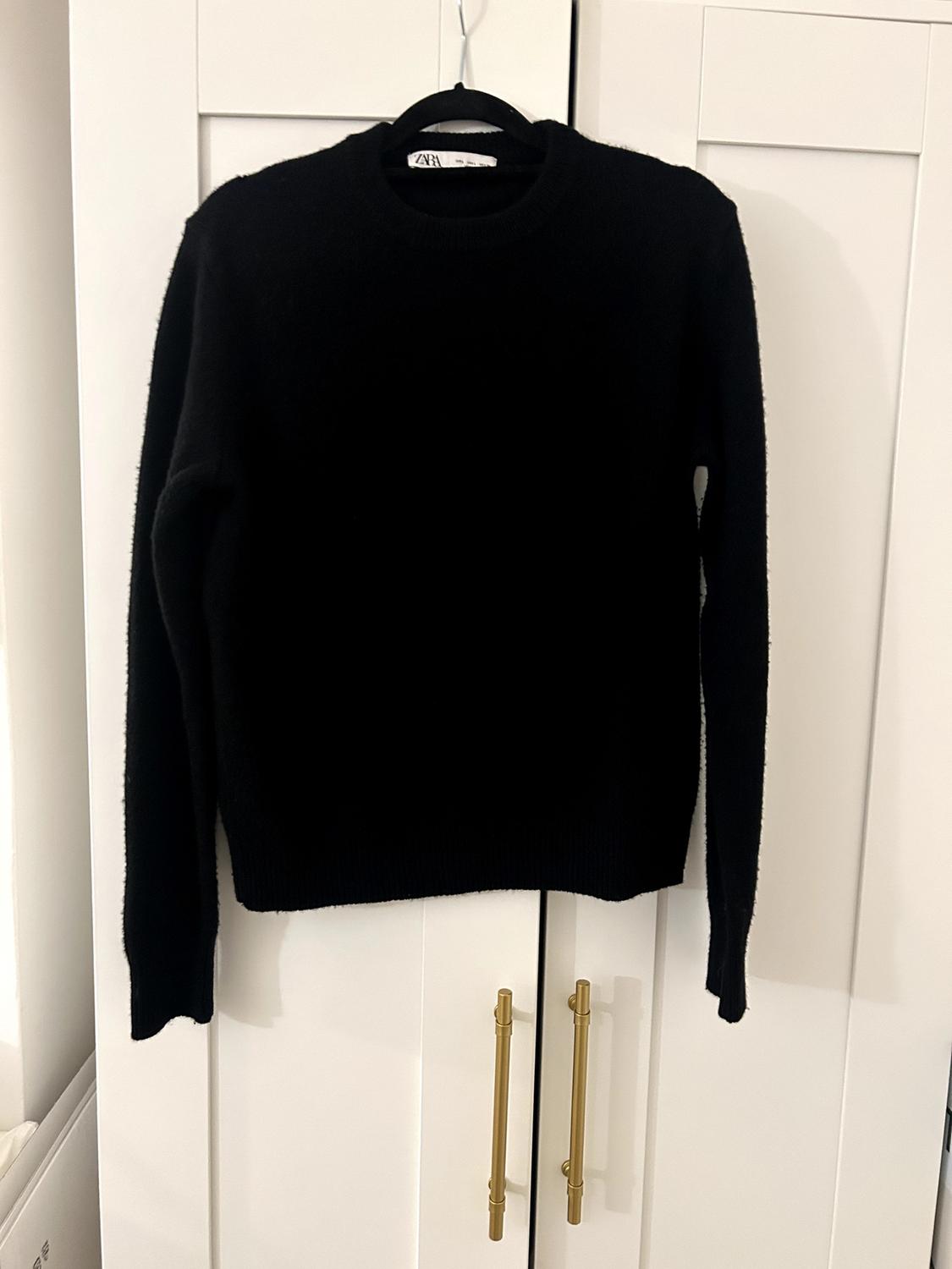 Black jumper hanging on a hanger in front of a white door with gold handles. Visible label with "ZARA" text.
