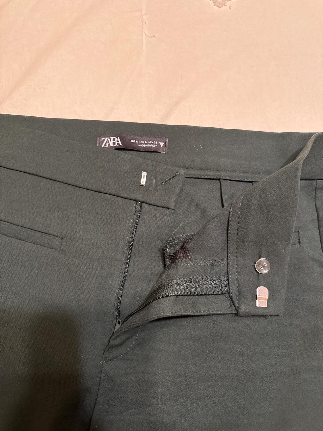 Dark trousers with open zip and visible button. Inner label with text: "ZARA EUR M USA M MEX 28 MADE IN TURKEY". Beige background.