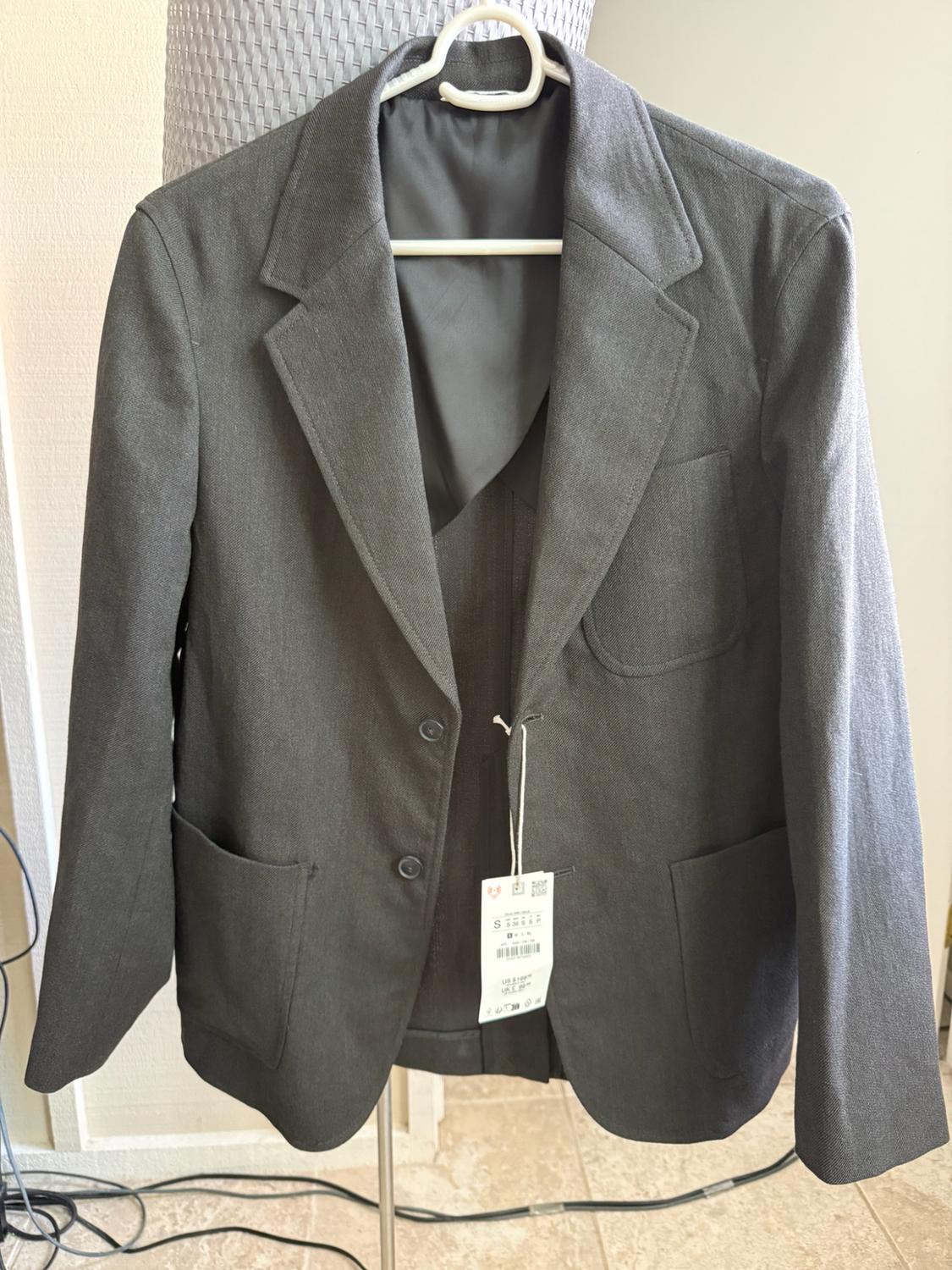 Dark gray two-button blazer with patch pockets, hanging on a white hanger indoors. It retains a white tag.