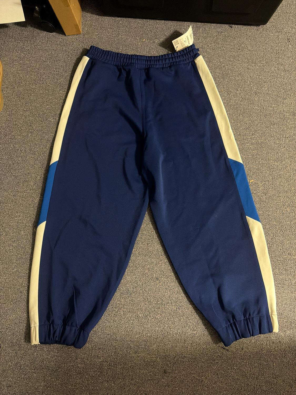 Dark blue tracksuit bottoms with white and blue side stripes, elasticated waist and cuffs, laid out on a grey carpet.