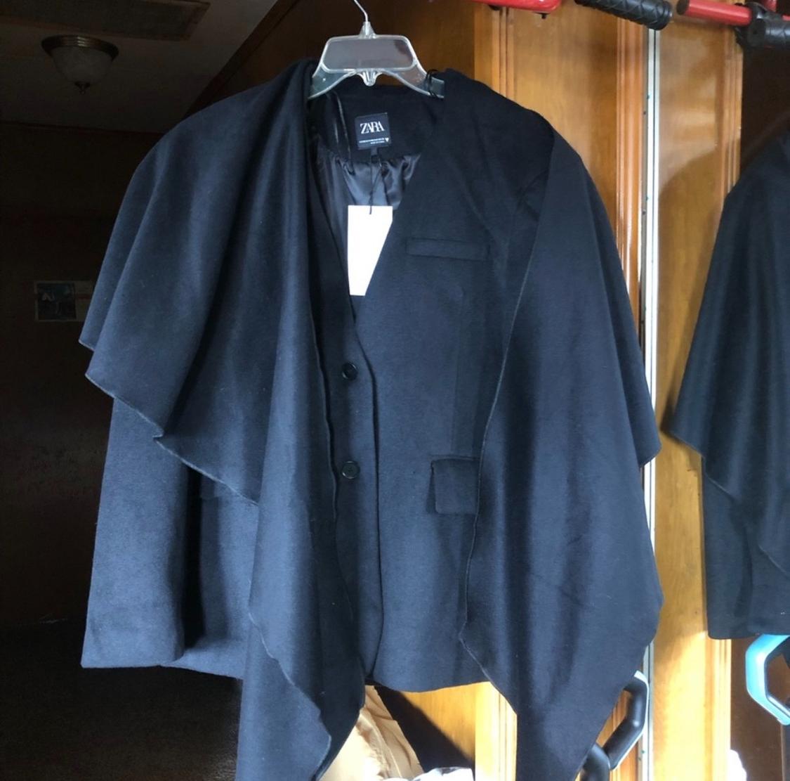 Black cape hanging on a hanger with a visible Zara tag, against a wooden background.