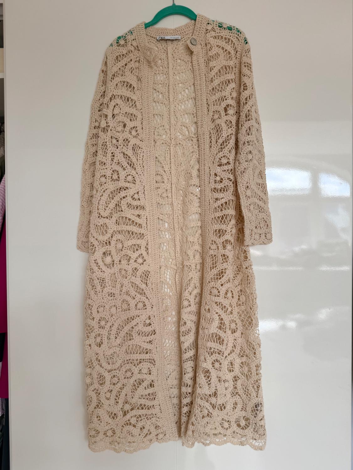 Long beige open-knit cardigan with long sleeves, hanging on a green hanger over a white door.