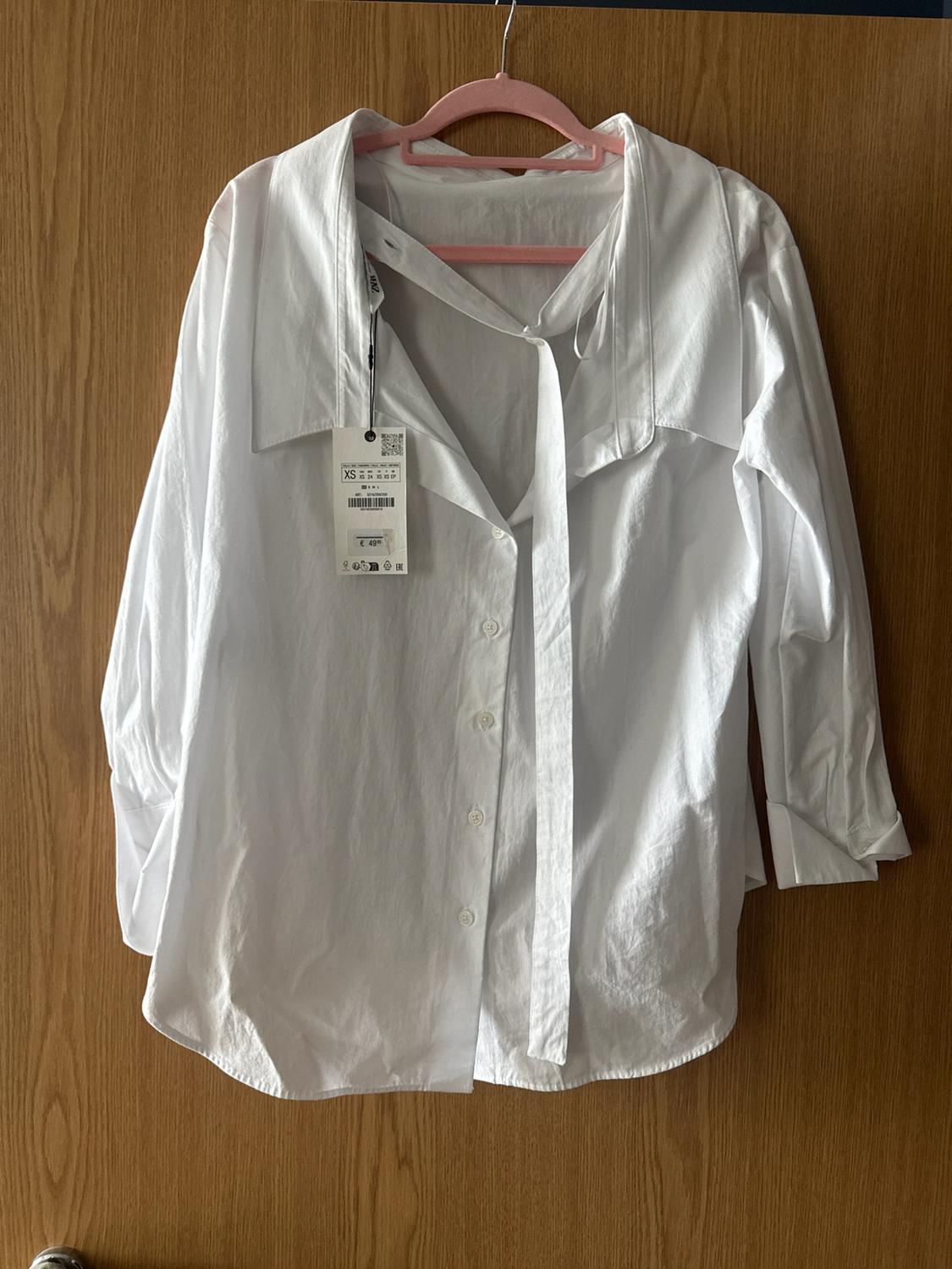 Long-sleeved white button-up shirt with a bow, hanging on a pink hanger on a wooden door. Label with the text XS and €49.