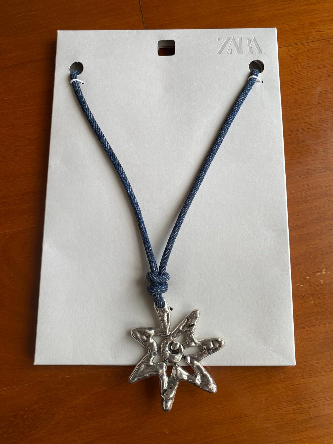 Blue cord necklace with a silver star-shaped pendant on white card bearing the text ZARA, on a wooden table.