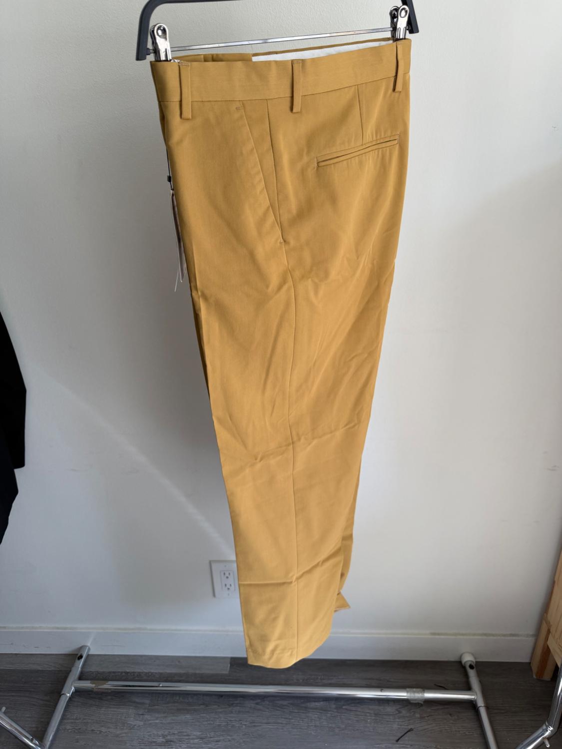 Mustard yellow trousers hanging on a metal hanger against a white background. Grey wooden floor visible at the bottom.