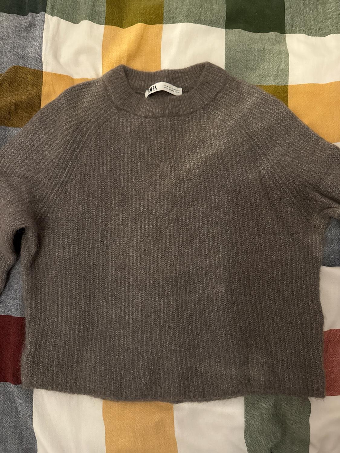 Grey crew neck knitted jumper with a ZARA label, on a multicoloured check fabric background.