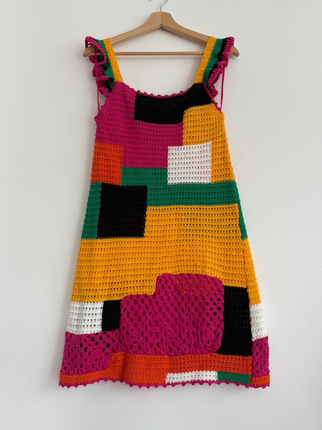 Multicoloured crochet dress with blocks in pink, orange, green, black and white, hanging on a wooden hanger against a white background.