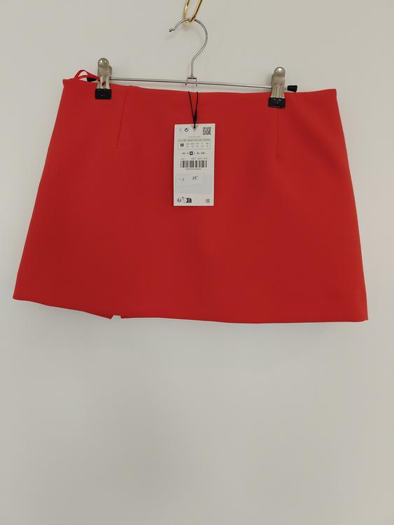 Red mini skirt hanging on a hanger with clips, visible label with size and price details on a white background.