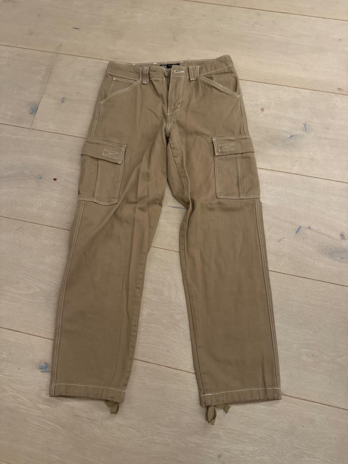 Beige cargo trousers with side pockets, laid on a light wooden floor.
