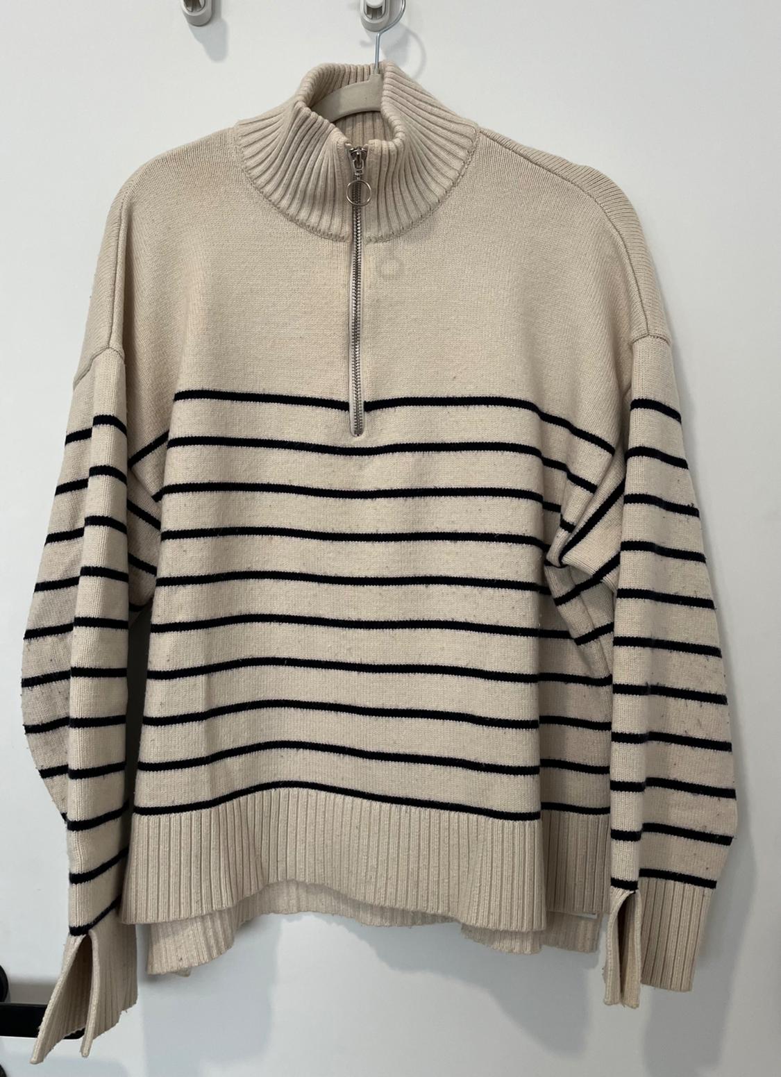 Beige jumper with black stripes, hanging on a hanger, with a high neck and front zip. White background.