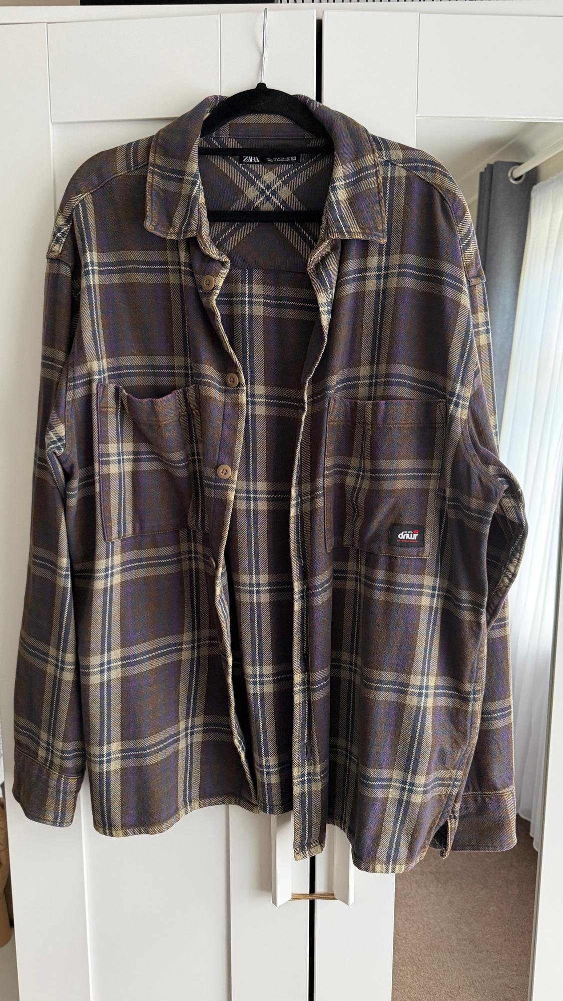 Brown and beige checked shirt hanging on a black hanger. Visible label: "Zara". Front flap pockets.