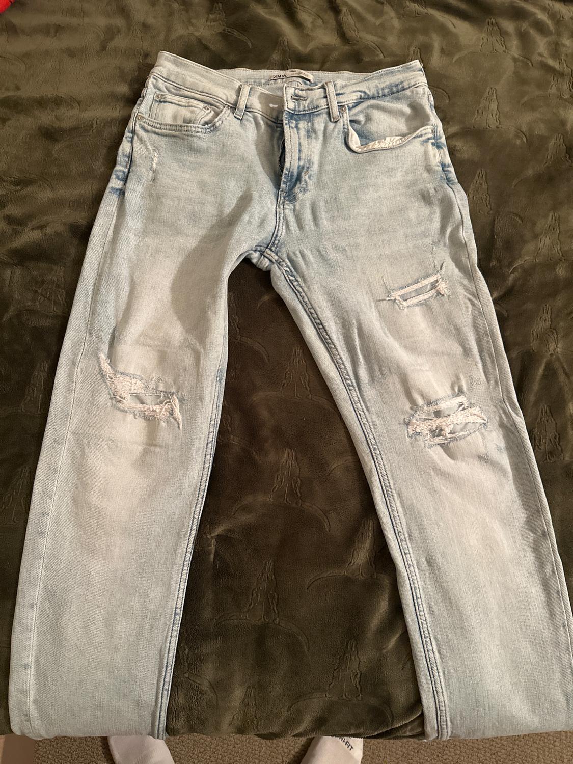 Light blue ripped-knee jeans, laid out on an olive green blanket. Label with the text ZARA.