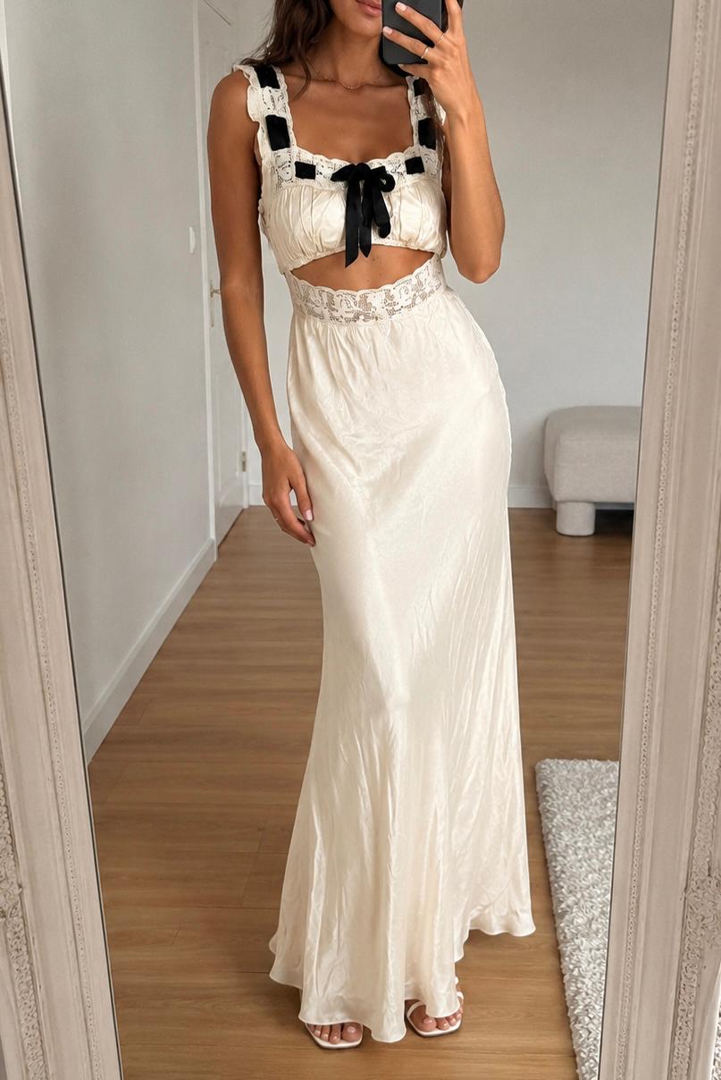 Woman taking a mirror selfie in a cream lace-trim top with black bows, a long satin skirt, and white sandals.