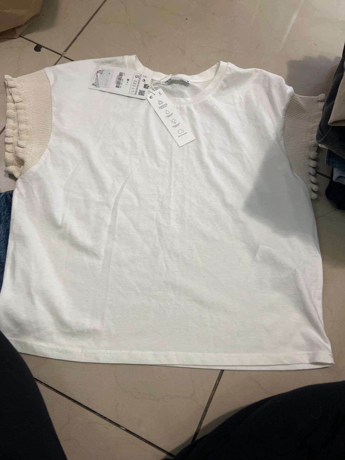 White T-shirt with short, beige fabric sleeves and pom-pom detailing. Visible tags on the garment. Light tiled background.