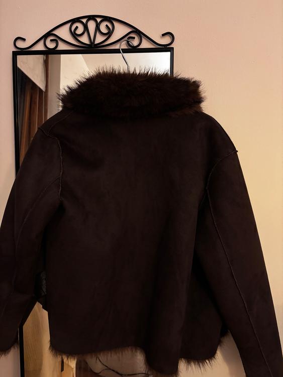 Brown jacket with a fur collar hanging on a mirror with a decorative black frame on a beige wall.