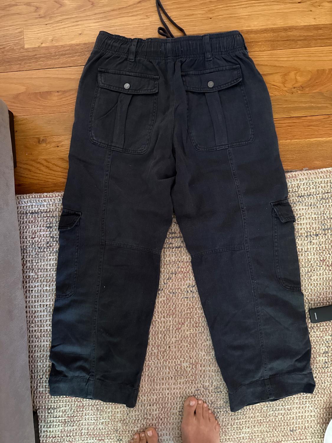 Black cargo pants with back and side pockets, on a beige rug. Bottom of feet visible in the image.
