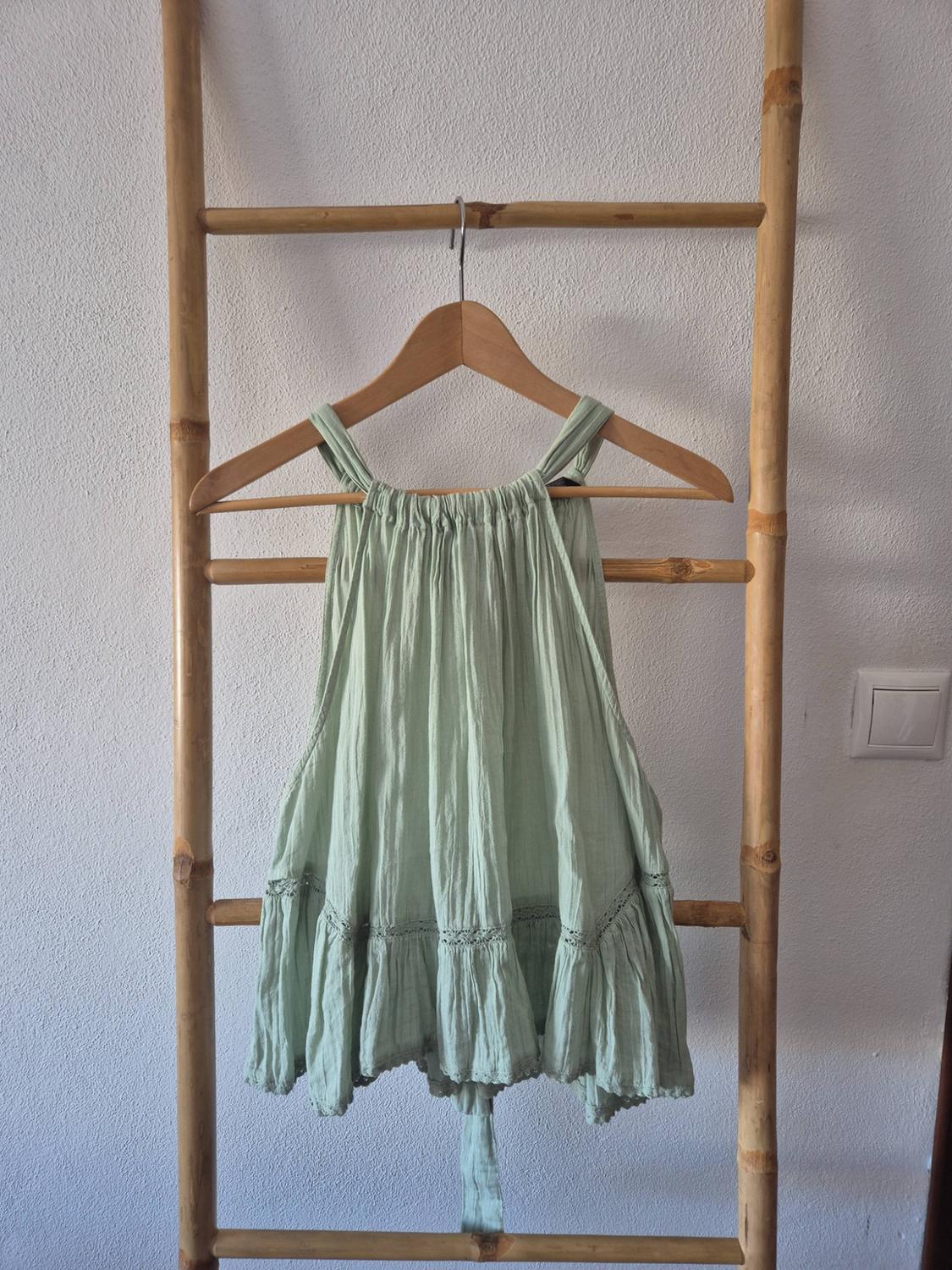 Light green halterneck blouse with gathered detailing and ruffles, hanging on a wooden hanger over a bamboo ladder against a white wall.