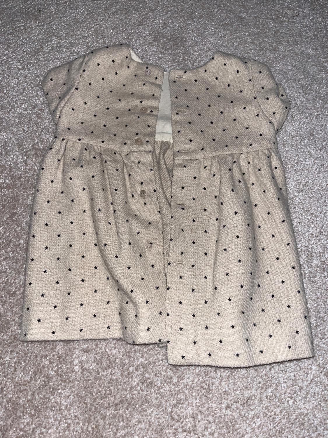 Beige baby dress with small black star print, short sleeves and buttons on the back, on a grey rug.
