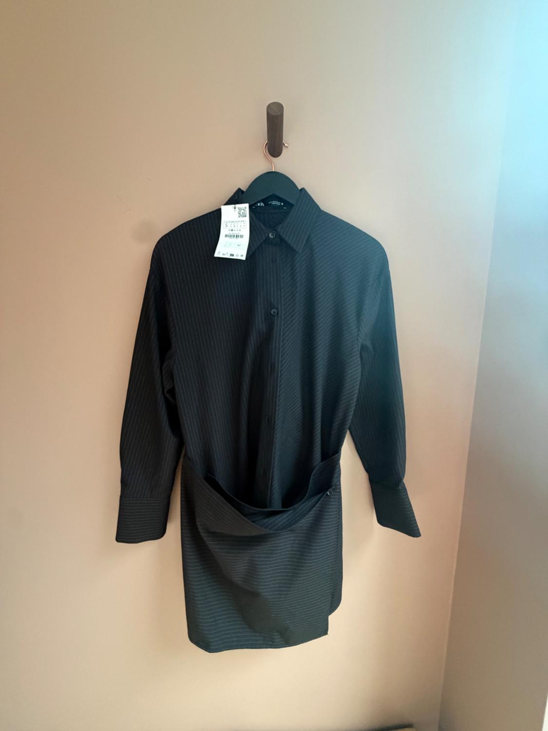 Black long-sleeved shirt dress with fine stripes, hanging on a plain wall. It stands out for its draped detail at the front.