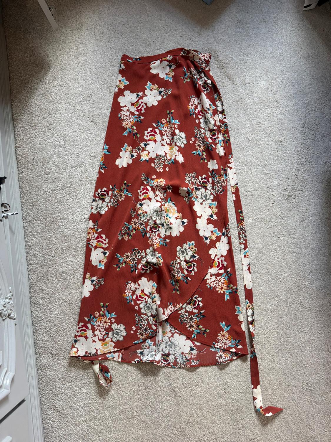 Long wrap-around skirt in terracotta with a floral print and side tie, laid out on a grey rug next to a white piece of furniture.