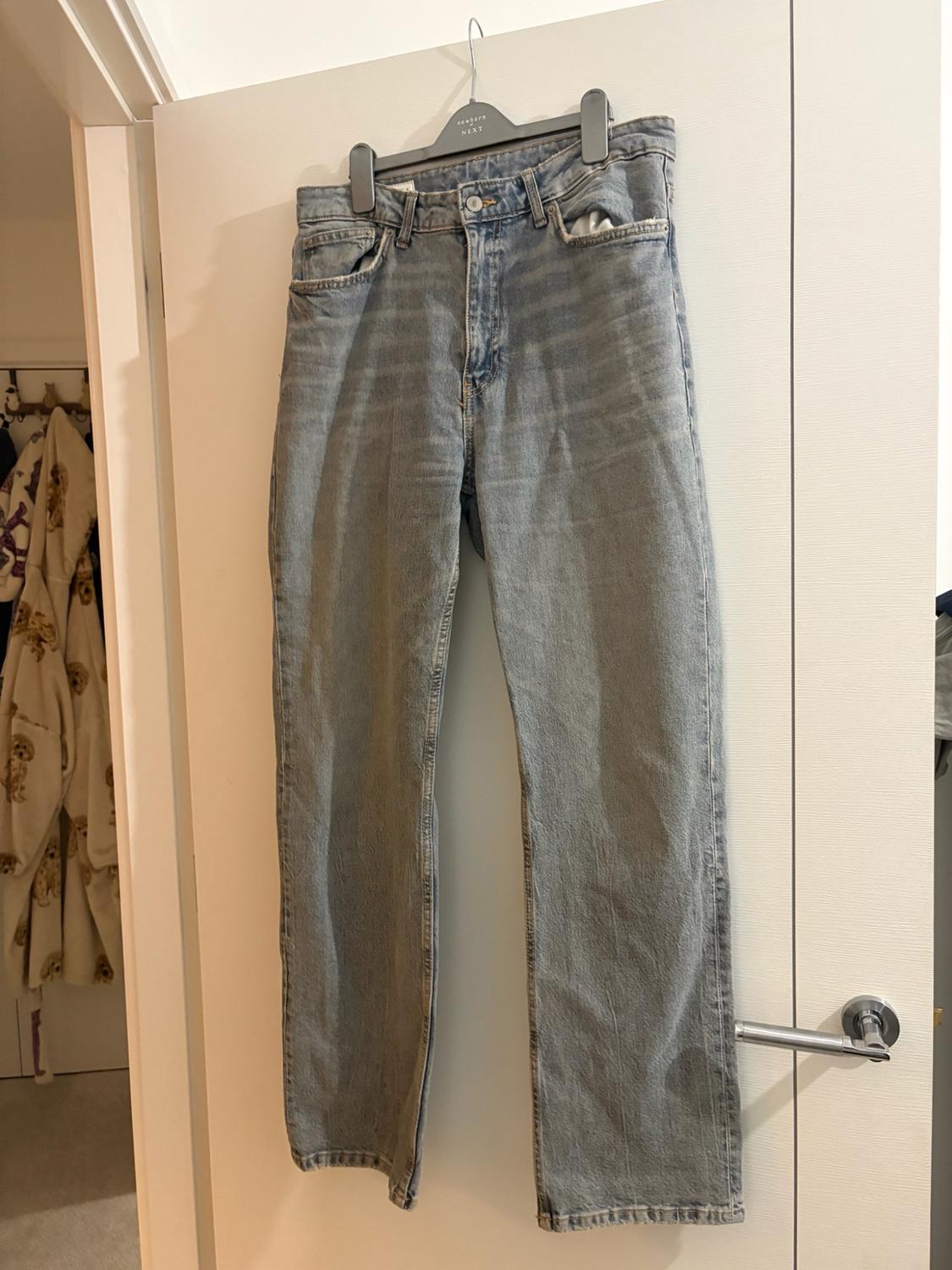 Light blue jeans hanging on a white door. Text on the hanger: newborn NEXT. In the background, you can see a room.