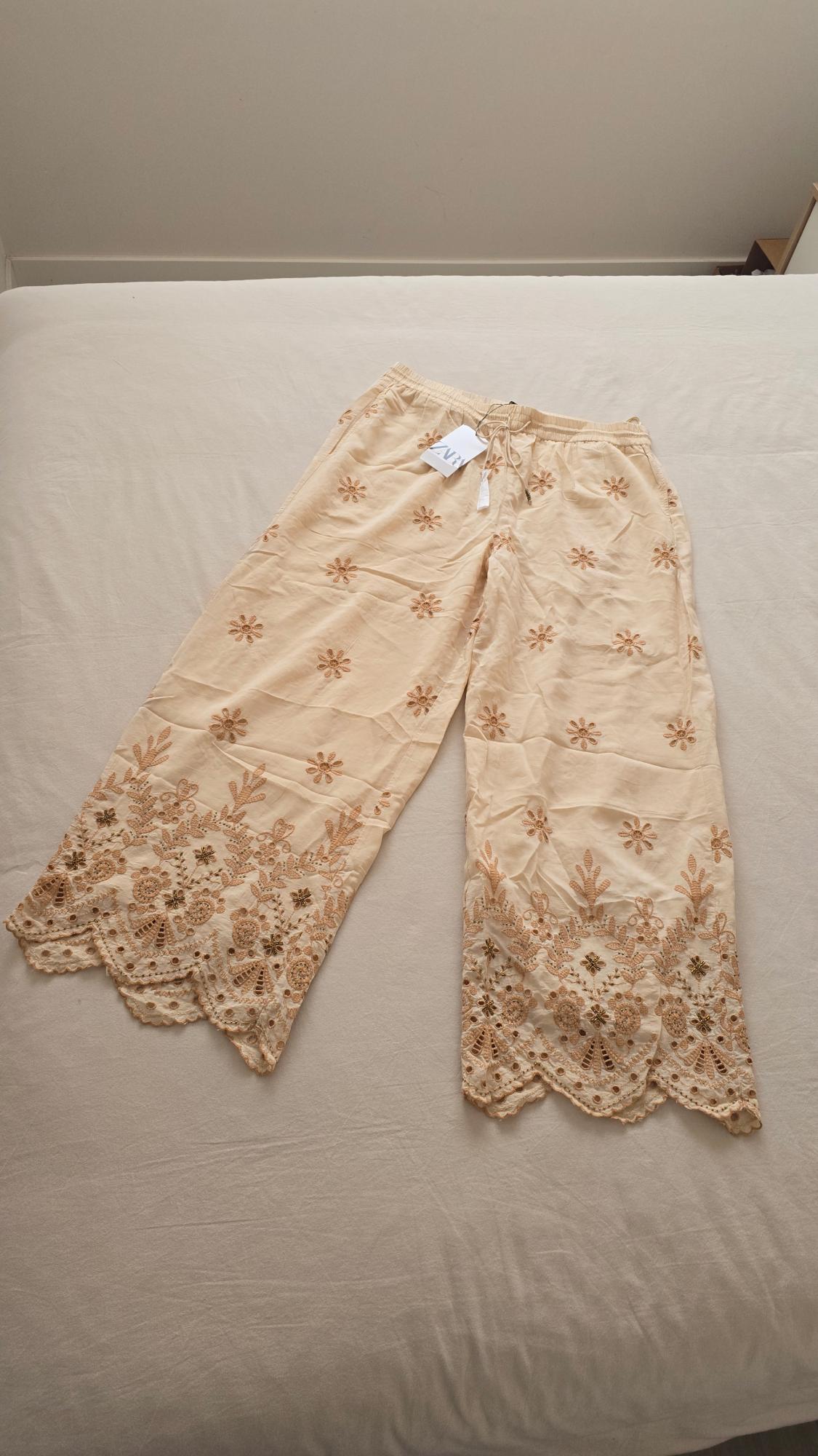 Beige trousers with brown floral embroidery and lace trim, on a white bed. Visible label with text "ZARA".