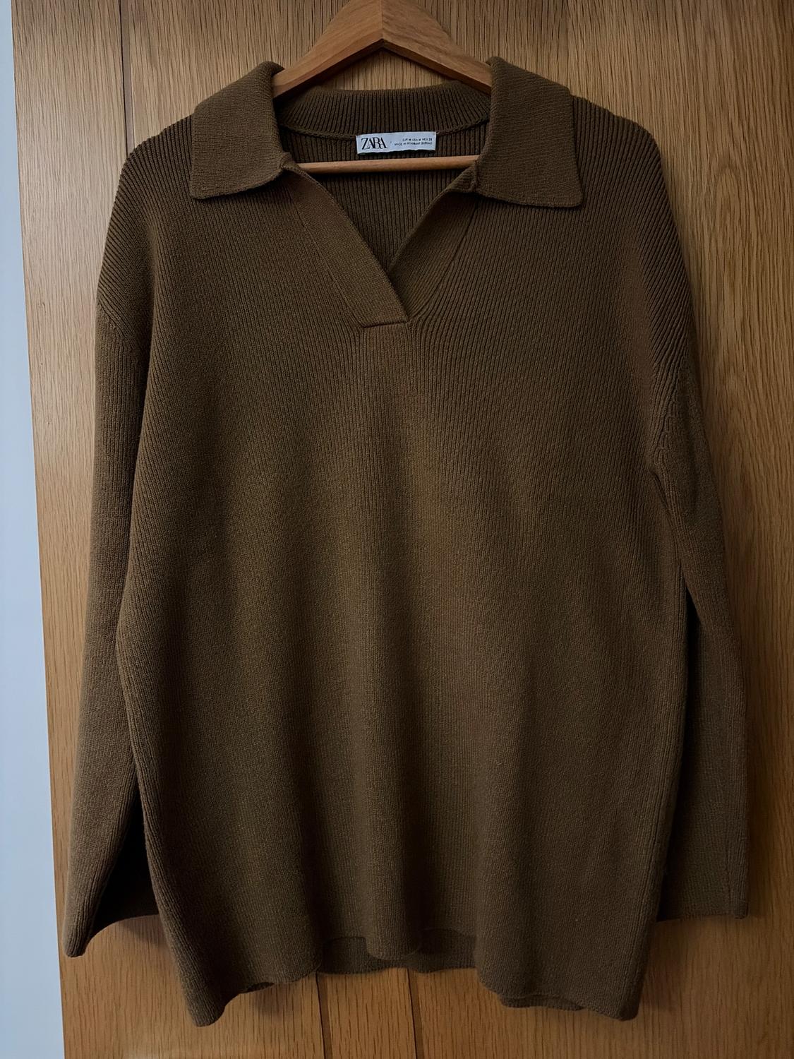 Brown knitted jumper with an open collar, hanging on a wooden hanger over a wooden door. Visible label: "ZARA".