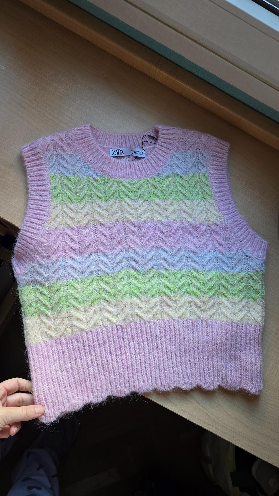 Sleeveless knitted vest in pastel pink, green, and beige tones with a zigzag pattern, on a wooden table. Visible label: ZARA.