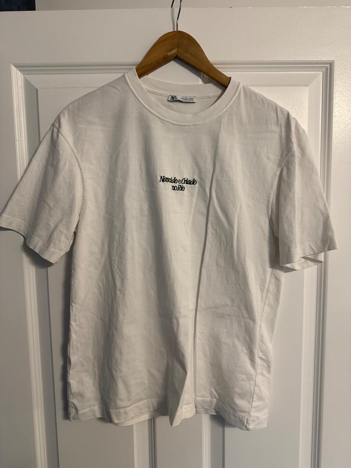 White t-shirt hanging on a wooden hanger, with embroidered text on the chest that reads "Born and raised in Rio". White door in the background.