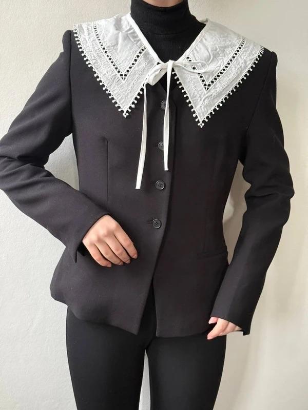 Model wearing a black button-up jacket with a white lace collar, over a black roll-neck jumper. White background.