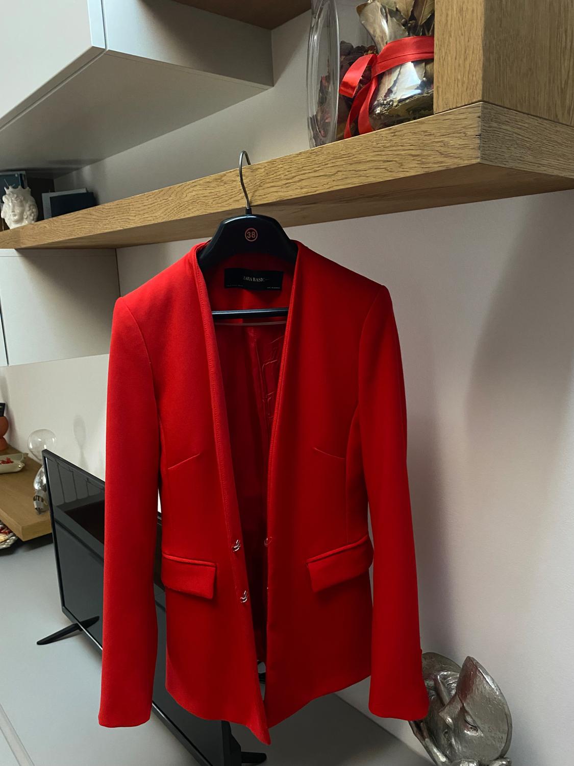 Red jacket hanging on a black hanger, against a backdrop of a wooden bookshelf with decorations and a white wall.
