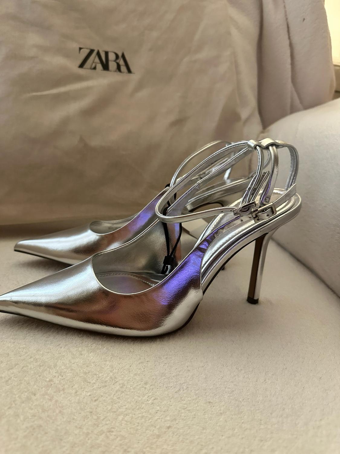 Silver high-heeled shoes with straps and pointed toes on a soft surface. In the background, there is a bag bearing the text ZARA.