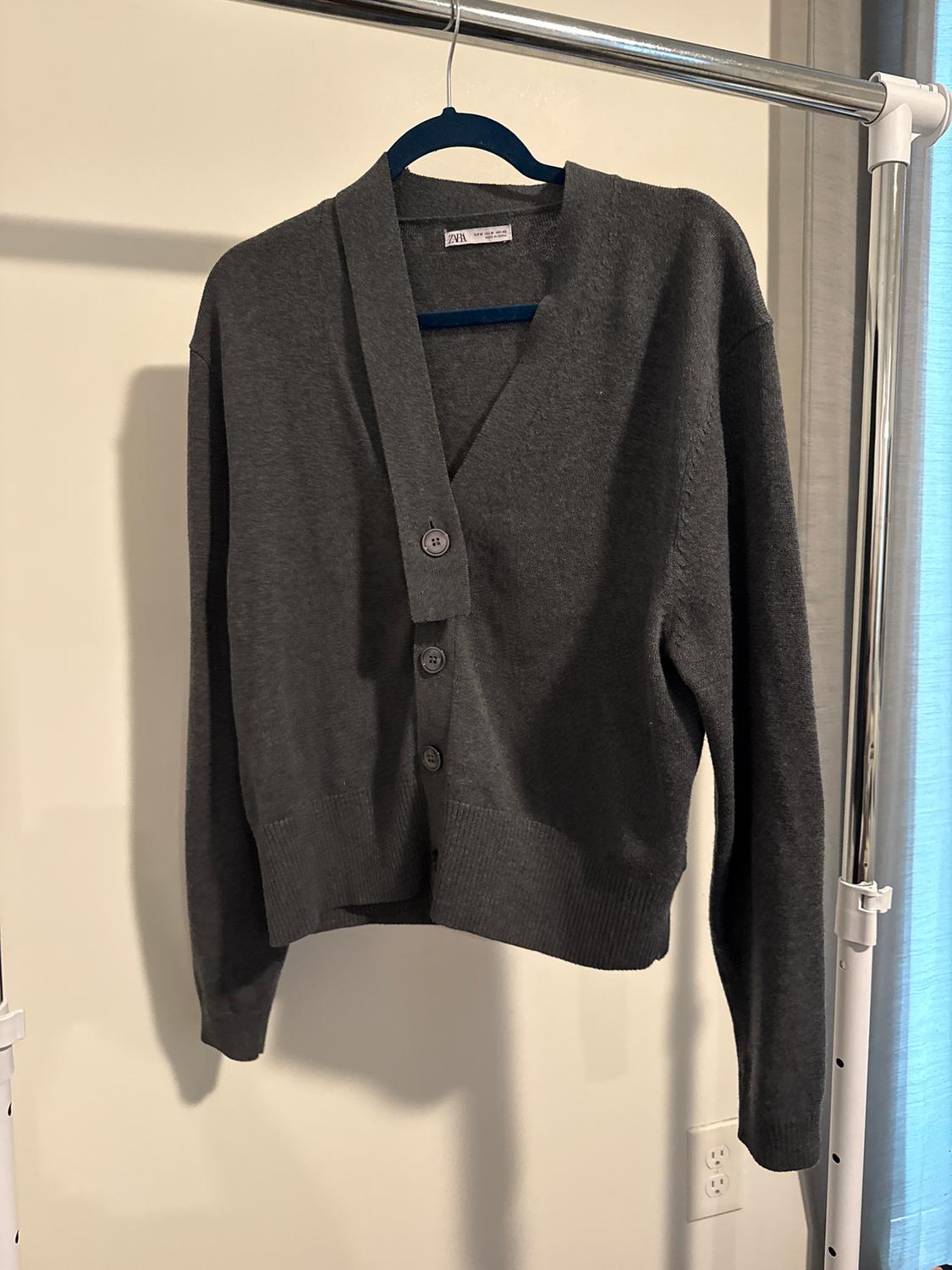 Grey knitted cardigan hanging on a blue hanger on a metal stand, with large buttons and a V-neck.