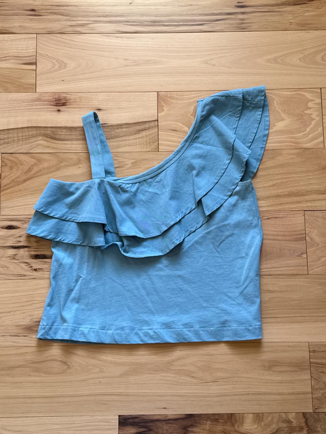 Light blue one-shoulder top with asymmetrical ruffle design, placed on a light wood floor with visible grain.