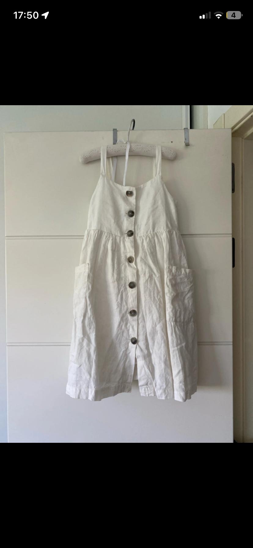 White strappy dress with front buttons and pockets, hanging on a hanger on a white door. Casual and relaxed style.