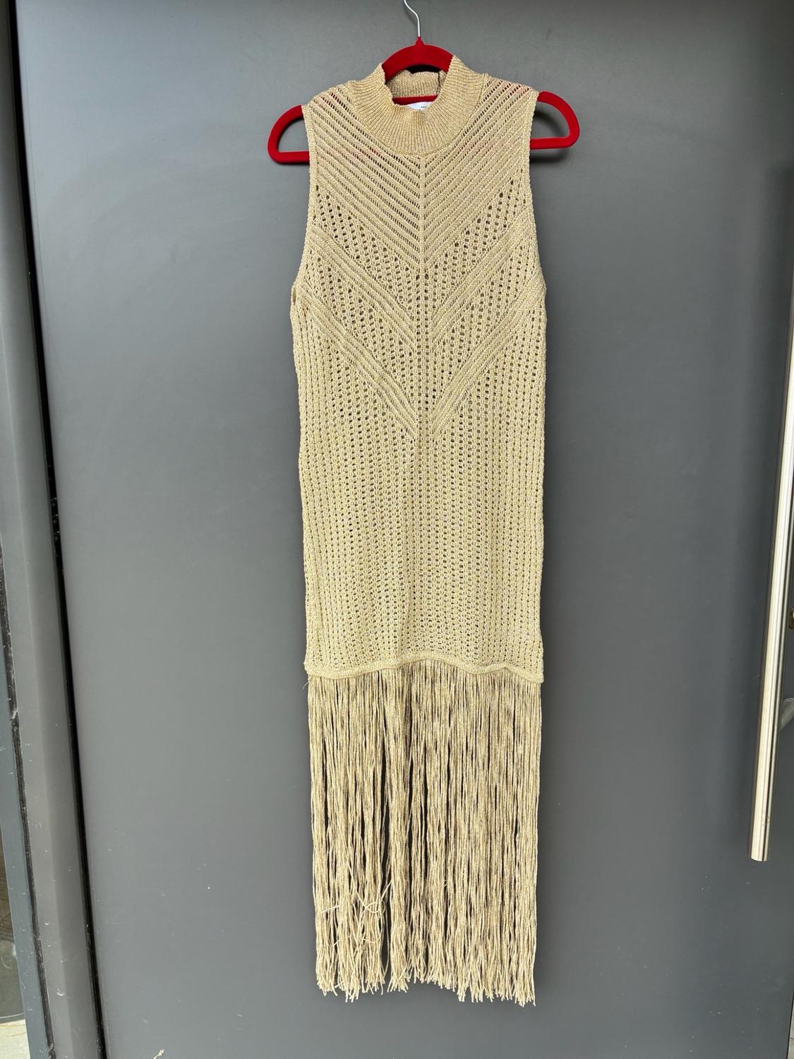Beige αμάνικo πλεκτό φόρεμα με ψηλό λαιμό και μακριά κρόσσια, κρεμασμένο σε μια κόκκινη κρεμάστρα σε μια γκρίζα πόρτα.