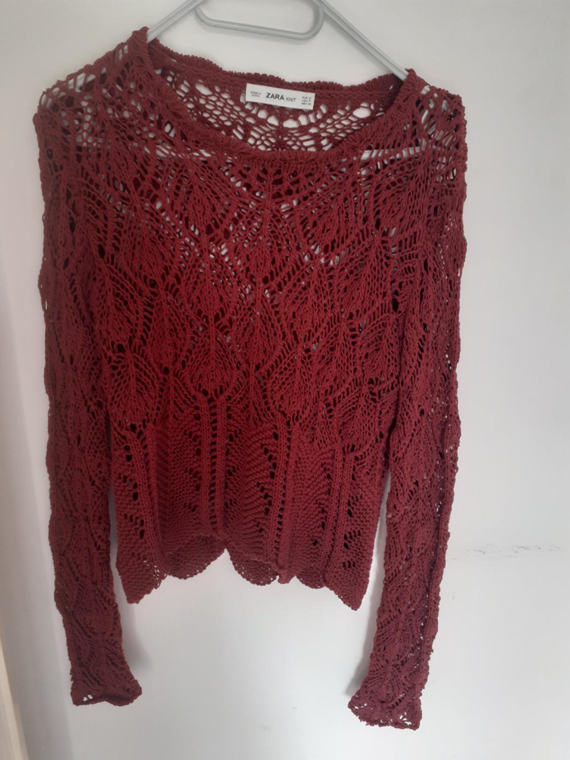 Maroon open-knit jumper with a round neck and long sleeves, hanging on a hanger against a white wall. Label with the text ZARA KNIT.