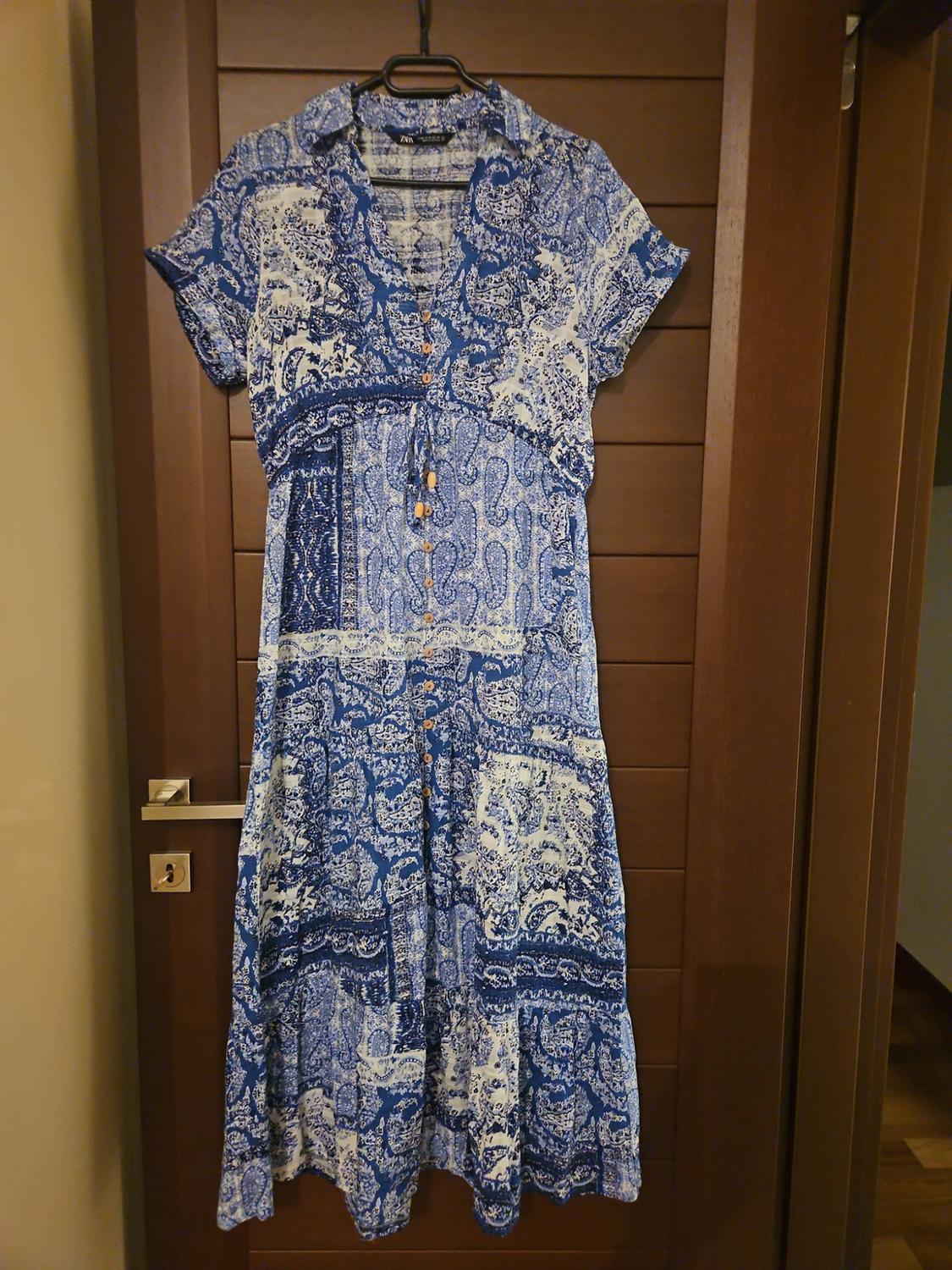 Blue and white patterned maxi dress, short sleeves, button-through with a drawstring waist, hanging on a wooden door. ZARA label.