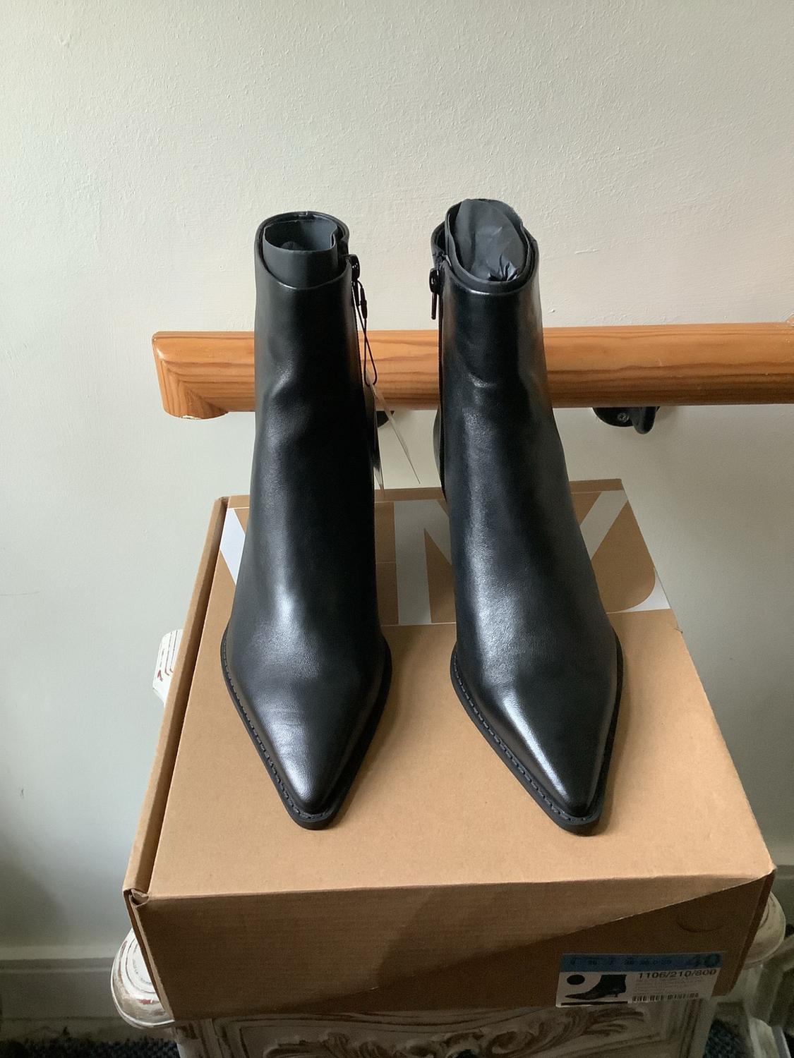 Black pointed-toe ankle boots on a cardboard box bearing the text NY, 40 and 1106/210/800. Background with a white wall and wooden handrail.
