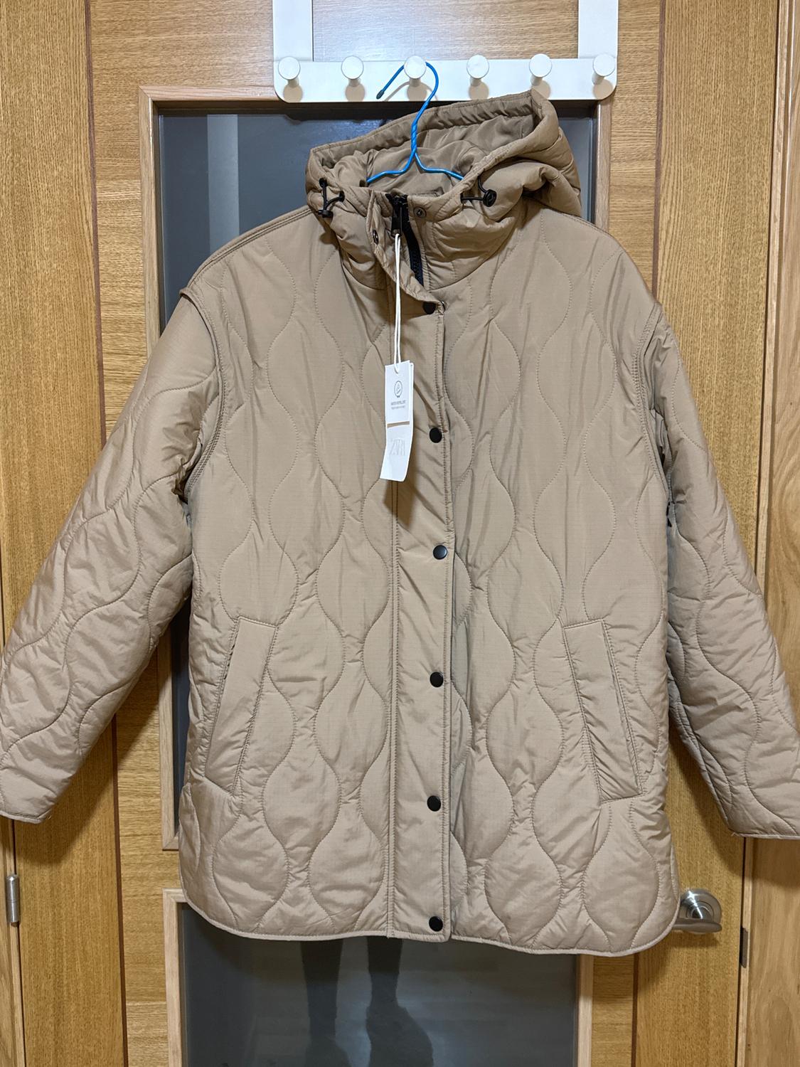 Beige padded jacket hanging on a wooden door. It has a hood, black buttons, and a white tag hanging from the collar.