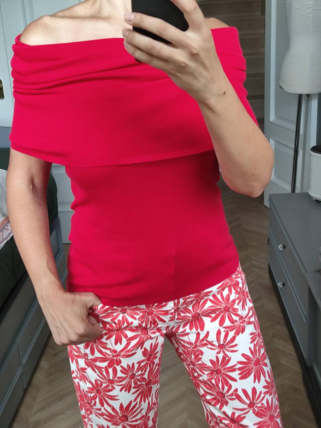 Person in front of a mirror taking a photo, wearing a red off-the-shoulder top and white trousers with a red floral print.