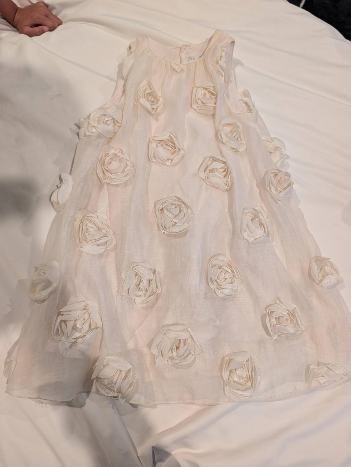 Light-coloured children’s dress made of translucent fabric with rose appliqués, on a white sheet. ZARA label visible at the neck.